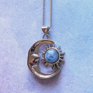 May include: Silver pendant necklace with a crescent moon and sun design. The sun features a round, light blue stone. The necklace hangs on a delicate silver chain. The background is a soft, gradient of blue and purple.