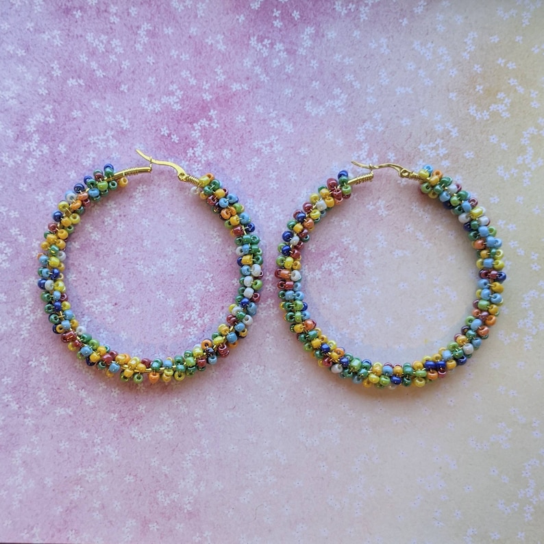 May include: A pair of large hoop earrings made with colorful seed beads. The beads are arranged in a spiral pattern and are a mix of green, blue, yellow, red, and white.