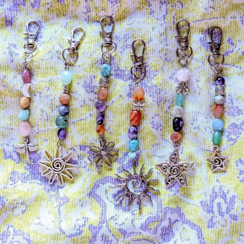 May include: Six silver-toned metal keychains featuring assorted colorful gemstone beads and charms. The charms include a dragonfly, a sun, a flower, and a star. Each keychain is attached to a lobster clasp, showcasing various natural stone shapes.