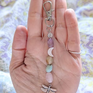 May include: A silver-toned metal keychain featuring a lobster clasp, a small flower charm, a white crescent moon charm, and a series of smooth, polished, multi-colored oval stones in shades of purple, tan, blue, and pink, ending with a dragonfly charm.