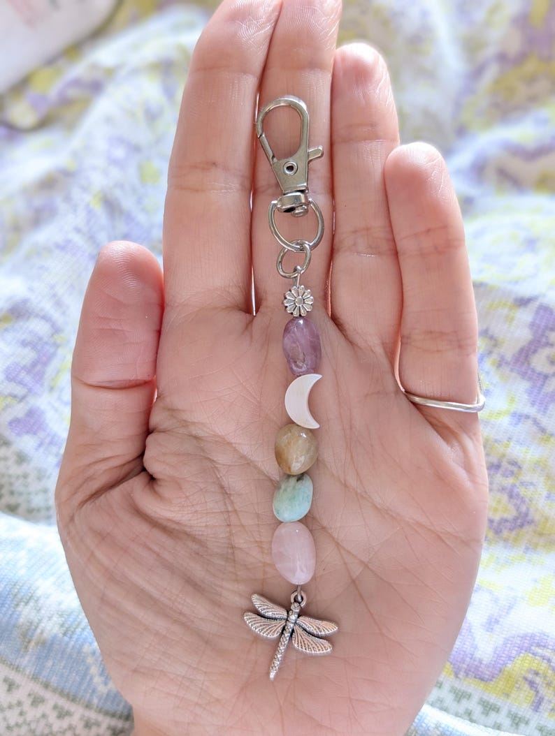 May include: A silver-toned metal keychain featuring a lobster clasp, a small flower charm, a white crescent moon charm, and a series of smooth, polished, multi-colored oval stones in shades of purple, tan, blue, and pink, ending with a dragonfly charm.
