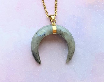 Könnte beinhalten: Halskette mit einem Anhänger in Form eines Halbmonds. Der Anhänger ist ein hellgrauer Stein mit dunkelgrauen und schwarzen Einschlüssen. Er hängt an einer goldfarbenen Kette. Der Anhänger ist mit einem goldfarbenen Verschluss an der Kette befestigt.