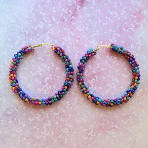 May include: A pair of large hoop earrings made of colorful beads. The beads are arranged in a spiral pattern and are in shades of blue, green, orange, pink, and purple. The earrings are made with gold-toned hoops.