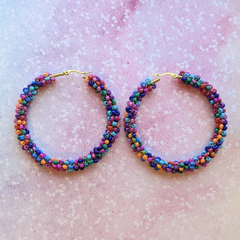 May include: A pair of large hoop earrings made of colorful beads. The beads are arranged in a spiral pattern and are in shades of blue, green, orange, pink, and purple. The earrings are made with gold-toned hoops.