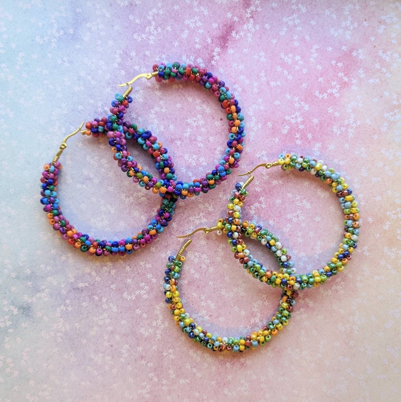 May include: Three pairs of colorful beaded hoop earrings. The hoops are made of small, colorful beads in a variety of shades, including pink, blue, green, yellow, and orange. The hoops are all different sizes, with the largest hoop being in the center of the image.