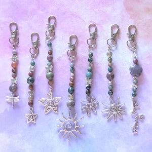 May include: Seven beaded keychains featuring silver-toned lobster clasps and various charms including a dragonfly, stars, a sun, a flower, and a leafy branch. The beads are multi-colored, featuring shades of green, brown, and grey, with silver accents.