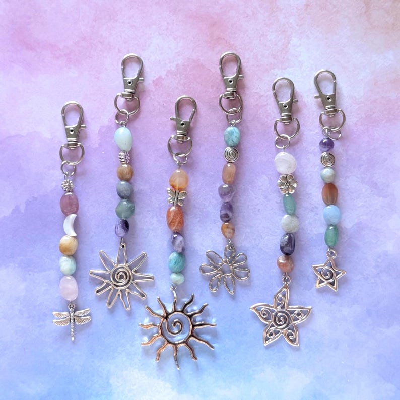 May include: Six silver-toned metal keychains featuring assorted polished gemstone beads in shades of pink, blue, purple, and orange. Each keychain includes a lobster clasp and a unique charm, including a dragonfly, suns, a flower, and a star.