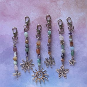 May include: Six silver-toned metal keychains featuring lobster claw clasps, assorted colorful polished gemstone beads, and dangling charms including a dragonfly, two suns, a flower, and a star, arranged vertically on a light purple background.