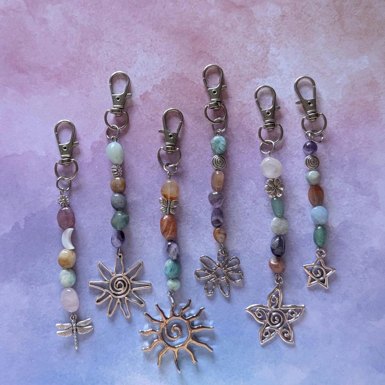 May include: Six silver-toned metal keychains featuring lobster claw clasps, assorted colorful polished gemstone beads, and dangling charms including a dragonfly, two suns, a flower, and a star, arranged vertically on a light purple background.
