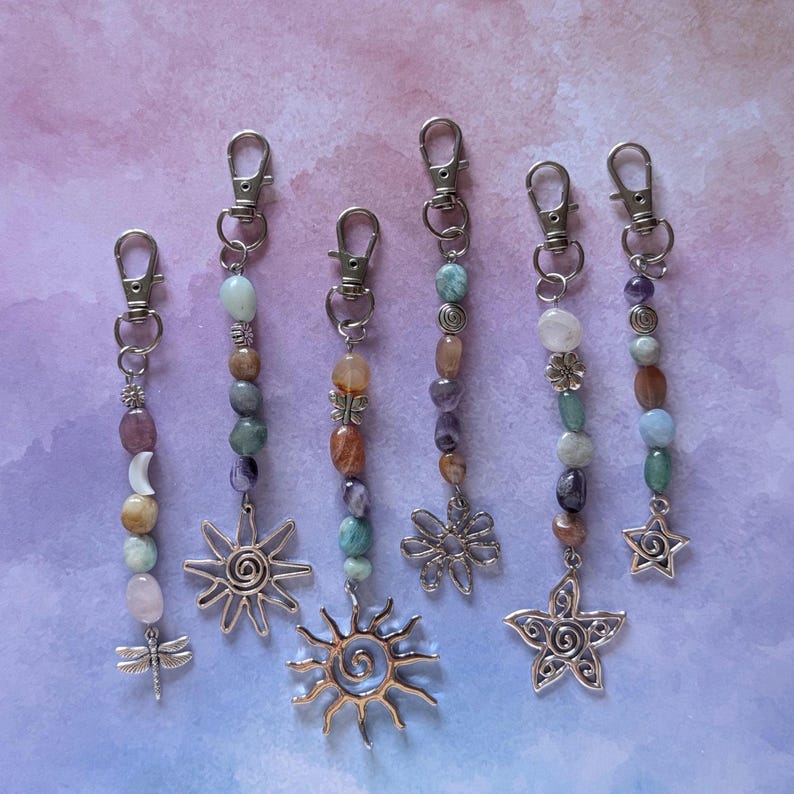 May include: Six silver-toned metal keychains featuring assorted polished gemstone beads in shades of pink, blue, green, and brown. Each keychain includes a lobster clasp and a unique charm, including a dragonfly, suns, flowers, and a star.