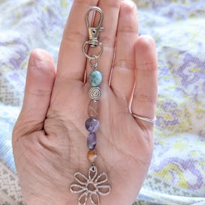May include: A silver-toned metal keychain featuring a lobster clasp, a small silver spiral bead, and a series of smooth, multi-colored polished stones in shades of blue, purple, and brown, ending with a silver-toned metal flower-shaped charm.