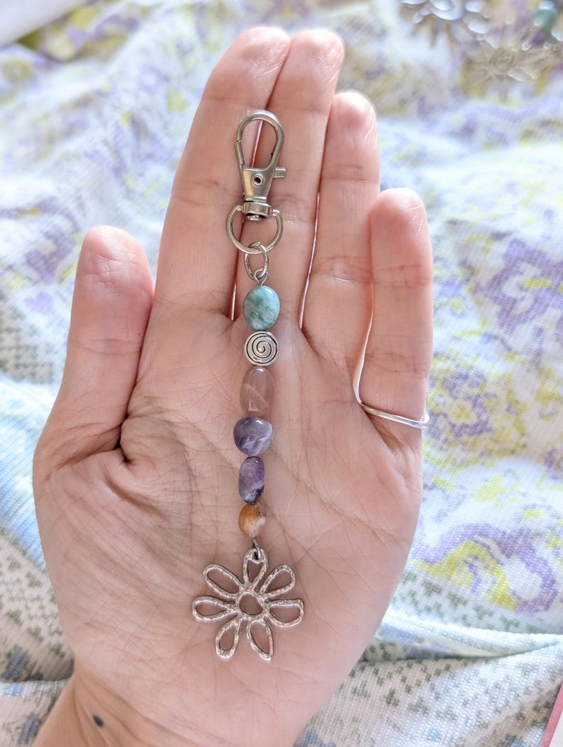 May include: A silver-toned metal keychain featuring a lobster clasp, a small silver spiral bead, and a series of smooth, multi-colored polished stones in shades of blue, purple, and brown, ending with a silver-toned metal flower-shaped charm.