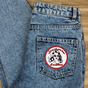 Può includere: Una tasca di jeans blu con una toppa ricamata in rosso e bianco che dice "Lasciami entrare in casa tua, sono una femminista".