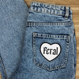 Op de afbeelding: Een paar blauwe denim jeans met een witte hartvormige patch waarop in zwarte gotische letters "Feral" staat.