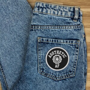 May include: A pair of blue denim jeans with a black and white patch on the back pocket that says "Aesthetic AF" with a peach graphic inside a circle.