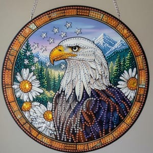 May include: A circular wall hanging featuring a diamond art depiction of a bald eagle in profile. The eagle is surrounded by white daisies, a pine forest, and snow-capped mountains under a sky with white stars, all framed by a decorative orange border.