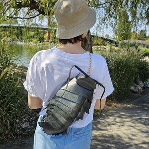 以下が含まれることがあります： ベージュのバケットハットと白いTシャツを着た人が、黒いセグメント状の甲虫の形をしたバックパックを背負っています。