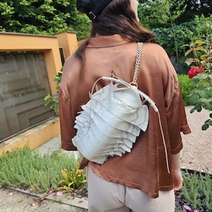 Può includere: Uno zaino bianco a strati con una tracolla a catena e un design unico e strutturato. La borsa è indossata sopra una camicia di raso marrone e pantaloni beige. Un berretto da baseball nero è indossato sulla testa.