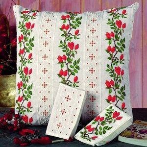 Puede incluir: Fundas de cojín blancas bordadas con un diseño floral con bayas rojas y hojas verdes. Las fundas de cojín están sobre una superficie de madera con un libro y otros artículos decorativos.