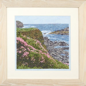 May include: A cross-stitch embroidery depicting a coastal scene with a rocky cliffside covered in pink flowers, overlooking a blue ocean with white waves crashing on the rocks.