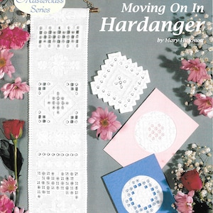 Könnte beinhalten: Ein weißes Hardanger-Stickprojekt mit dem Text "Moving On In Hardanger" und "Masterclass Series". Das Bild zeigt ein langes, rechteckiges Stück, eine quadratische Deckchen und zwei Karten, alle mit aufwendigen Stickmustern. Rosa Blumen und ein grauer Hintergrund vervollständigen die Szene.