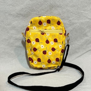 May include: Yellow crossbody bag with a ladybug and daisy pattern. The bag has a zippered top, a black adjustable strap, and a gold zipper. The bag measures 8" x 5.5" x 1.5".