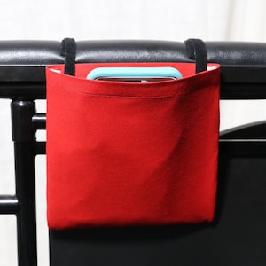May include: Red fabric pouch with black straps attached to the armrest of a black wheelchair. A light blue phone is inside the pouch.