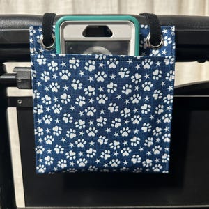 May include: A navy blue fabric organizer with a white paw print and star pattern, designed to hang on a mobility device. The organizer has a pocket holding a smartphone. Black loops and grommets secure the organizer.