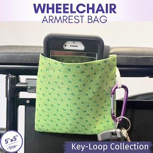 May include: A light green fabric wheelchair armrest bag with a small floral pattern, featuring a black smartphone inside the pocket, a white elastic loop, a purple carabiner, and a set of keys. The bag measures 5 inches by 5 inches.