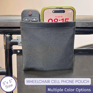 May include: A black fabric pouch measuring 5 inches by 5 inches, attached to a wheelchair armrest. The pouch contains a black remote control and a smartphone with a green case. Text on the image reads: WHEELCHAIR CELL PHONE POUCH, 5" x 5" Size.