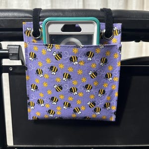 May include: A purple fabric pouch with a bee pattern hangs from the back of a chair. The pouch has a black strap and a clear plastic pocket for a phone or other device.