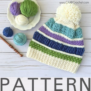May include: A white crochet hat with a pom pom on top. The hat has stripes of blue, green, purple, and white yarn. The hat is lying on a white wooden surface. The yarn balls are in the background. "PATTERN" is written in black letters at the bottom of the image.