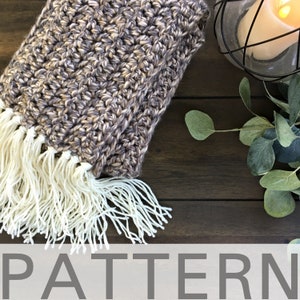 May include: A brown and beige crocheted blanket with a white fringe. The blanket is lying on a wooden surface with a candle holder and green leaves in the background. The text "PATTERN" is printed in gray at the bottom of the image.
