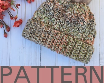 Op de afbeelding: Een gebreide muts met een bruin, groen en oranje kleurpatroon. De muts heeft een nep bont pompon bovenop. De muts ligt op een wit houten oppervlak. De tekst "PATTERN" staat onderaan de afbeelding.