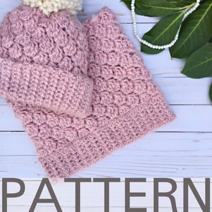 May include: A pink crocheted hat and matching neck warmer with a white pom pom on top. The set is laid out on a white wooden surface.