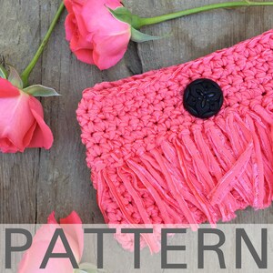 Könnte beinhalten: Eine rosa gehäkelte Clutch mit einem schwarzen Knopf und Fransen. Die Tasche liegt auf einer Holzoberfläche mit rosa Rosen.