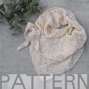 Puede incluir: Bufanda de cuello de ganchillo color crema con motas amarillas y azules. La bufanda está doblada y drapeada, mostrando su patrón de puntadas texturizadas. La palabra "PATTERN" es parcialmente visible en la parte inferior de la imagen.