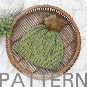 May include: A green knit beanie with a ribbed design and a brown faux fur pom-pom. The hat is displayed on a woven wicker tray. The word "PATTERN" is visible at the bottom of the image.