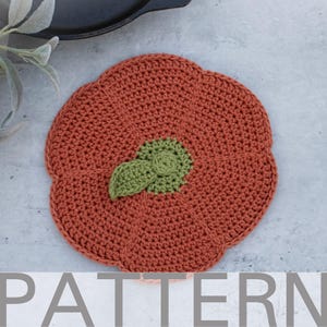 May include: A handmade, crocheted pumpkin-shaped trivet in shades of orange and green. The trivet features a detailed, textured design with a green leaf and stem accent in the centre. The word "PATTERN" is visible at the bottom of the image.