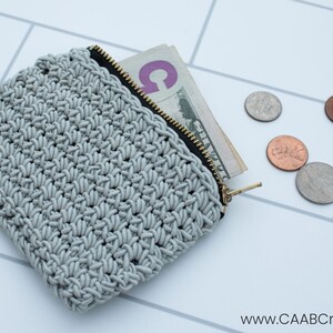 Puede incluir: Una bolsa de cremallera de crochet gris con una cremallera negra y algunos billetes de d&oacute;lar y monedas en su interior. La bolsa est&aacute; sobre una superficie de azulejos blancos.
