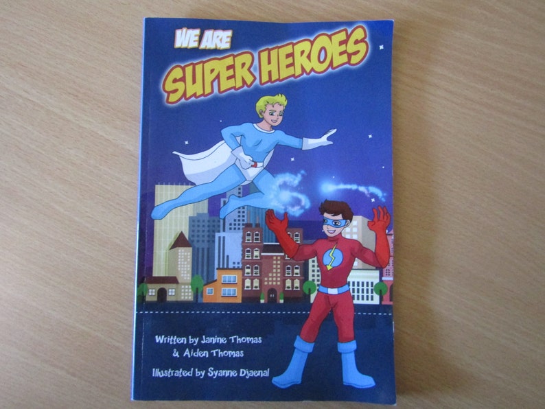 May include: Book cover with the title "We Are Super Heroes" in yellow letters on a blue background. Two superheroes, one in blue and white and the other in red, are flying in front of a city skyline. The book is written by Janine Thomas & Aiden Thomas and illustrated by Syanne Djaenal.