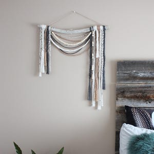 Peut inclure: Une décoration murale composée d'un morceau de bois flotté avec divers brins de corde en macramé, de corde tressée et de perles en bois de couleur blanche, beige et gris foncé. Les brins sont disposés en boucles drapées et en pompons.