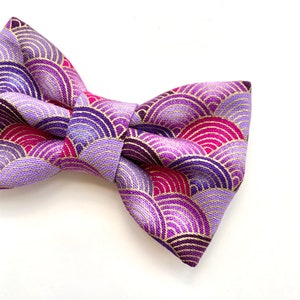 May include: A purple and pink bow tie with a gold geometric pattern. The bow tie is made of a soft, textured fabric.