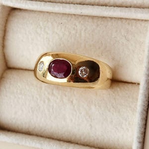 Op de afbeelding: Een gouden ring met een centrale ovaal geslepen rode robijn, geflankeerd door twee kleine rond geslepen heldere diamanten. De ring is gezet in een gepolijste geelgouden zetting met een breed, glad profiel, gepresenteerd in een crèmekleurig sieradendoosje.