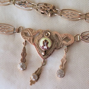 Op de afbeelding: Een goudkleurige ketting met een ingewikkeld ontwerp. De ketting heeft een centrale hanger met een miniatuurportret en bungelende accenten. De ketting heeft een opengewerkt patroon. De ketting wordt gepresenteerd op een witte ondergrond.