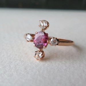 Pode incluir: Um anel de ouro rosa com uma gema rosa no centro e quatro gemas brancas menores ao redor.