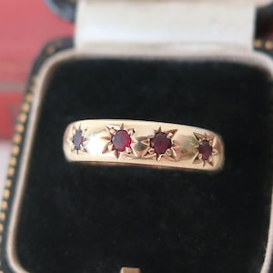 May include: A gold band ring featuring four small, round, deep red rubies set into the metal. Each ruby is surrounded by a star-shaped engraving that radiates outward from the stone, creating a distinct pattern across the surface of the gold band.