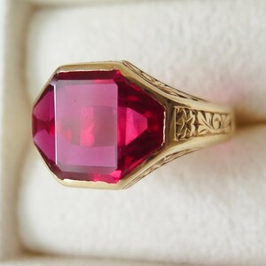 Pode incluir: Um anel de ouro com uma grande gema de rubi retangular facetada. O anel tem um design ornamentado e gravado na banda. O rubi é de um vermelho profundo e vibrante, e o anel é embutido em uma banda de ouro.