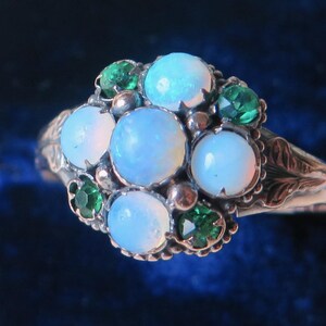 May include: A silver ring with a cluster of opalescent, round gemstones and smaller green stones. The ring's band has an ornate, floral design. The ring is set against a dark blue velvet background.