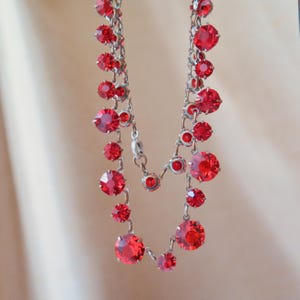 Puede incluir: Un collar deslumbrante con una serie de gemas rojas vibrantes de varios tamaños, unidas por una delicada cadena plateada. Las gemas, de corte redondo, brillan, creando un accesorio glamuroso. El collar es una pieza llamativa.